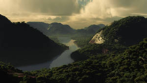 一條河流流經茂密的綠色山脈，天空多雲，帶有金色光芒。 — Calming 壁紙