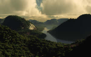 一條河流流經茂密的青山，天空多雲，帶有金色光芒。 — 平靜 壁紙