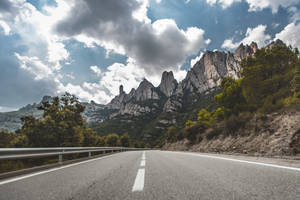 在多雲的天空下，蜿蜒的道路通往鋸齒狀的山脈。 — 山景 壁紙