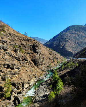 一條充滿活力的藍綠色河流流經岩石峽谷，兩旁是陡峭的懸崖和稀疏的樹木，在清澈的藍天下。 — 不丹 壁紙