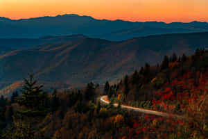 蜿蜒道路穿過秋日山脈，日落時分火紅橙色天空。 — MacBook Air M3 壁紙