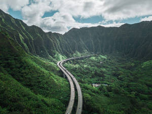 一條彎曲的高速公路在多雲的天空下穿過茂密的綠色山谷。 — 山脈 壁紙
