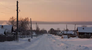 積雪的村莊道路，在光禿樹木和木屋之間，通往夕陽下的冰凍湖泊。 — 冬季風景 壁紙