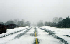 被雪覆蓋的道路，帶有黃色中心線，穿過霧氣瀰漫的冬季森林。 — 16:10 壁紙