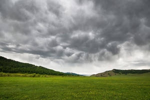 戲劇性暴風雲下的廣闊綠色草地，遠處有樹木繁茂的山丘。 — 風景 壁紙