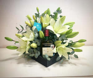 威士忌瓶被白百合和綠色植物環繞，放在黑色禮盒中。 — 花 壁紙