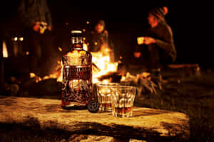 營火旁的威士忌酒瓶和酒杯，背景有人。 — 露營 壁紙