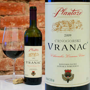 一瓶2009年Plantaže Vranac紅酒，附玻璃杯和軟木塞，放在桌上，背景是磚牆。 — 葡萄酒 壁紙