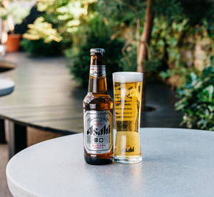 Asahi 啤酒瓶和玻璃杯放在露台桌上，背景有綠色植物。 — 啤酒 壁紙
