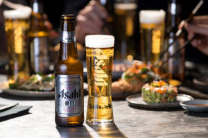 朝日啤酒瓶和玻璃杯，搭配壽司盤和筷子，在餐廳環境中。 — 日本 壁紙