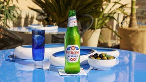 Peroni Nastro Azzurro 0.0 啤酒瓶、玻璃杯和橄欖在戶外藍色桌子上。 — 啤酒 壁紙