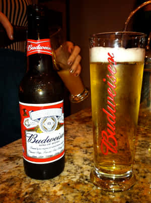 冰鎮的Budweiser啤酒瓶和玻璃杯放在大理石酒吧上，有一隻手拿著另一個玻璃杯。 — iPad Pro M4 壁紙