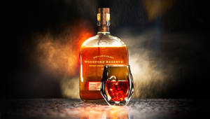 Woodford Reserve 波本威士忌瓶和帶冰塊的玻璃杯在酒吧上，有煙霧和溫暖的燈光。 — 煙動圖 壁紙