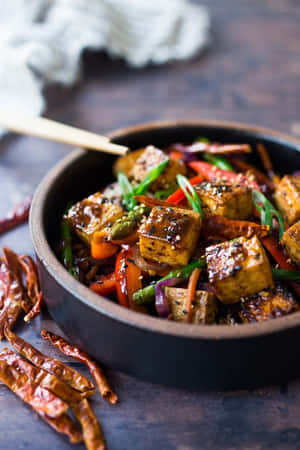 深色碗中的炒豆腐和蔬菜，旁邊有乾辣椒。 — 2:3 壁紙