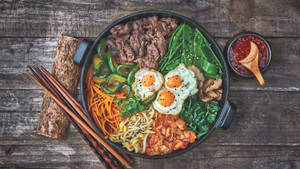 色彩繽紛的韓國石鍋拌飯，配有牛肉、雞蛋和蔬菜，放在木桌上，附有筷子。 — 烹飪 壁紙