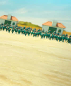 在沙地上兩座小建築物之間排成隊形的模糊軍事人員。 — 067 魷魚遊戲 壁紙