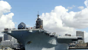 一艘大型灰色航空母艦停泊在城市港口，陽光明媚，甲板上有人。 — 德克薩斯州旗 壁紙