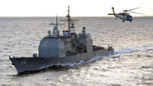 軍用驅逐艦在日落時分航行，直升機在海面上飛行。