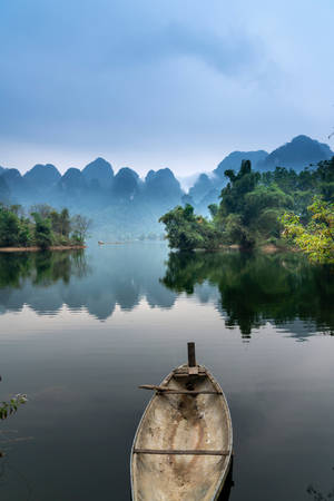 一艘小船在平靜的水面上休息，背景中霧氣繚繞的山巒倒映其中。 — 河景 壁紙
