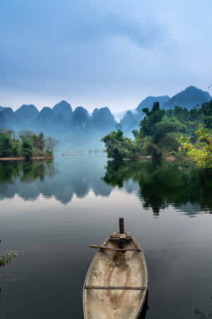 一艘小船停泊在平靜的河面上，四周環繞著霧氣繚繞的山脈和茂密的綠植。 — 小米平板 9 Pro Max 壁紙