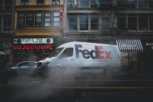 FedEx貨車在霧氣瀰漫的街道上行駛，兩旁有霓虹招牌和磚建築。 — 聯邦快遞 壁紙