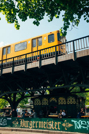 高架軌道上的黃色火車，下方是熙熙攘攘的戶外咖啡館，有人群和綠樹。 — 柏林 壁紙