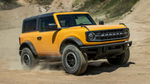 黃色福特Bronco SUV在塵土飛揚的沙漠地形上行駛。 — 福特 壁紙