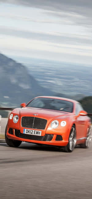 橙色 Bentley 跑車在模糊背景的山路上飛馳。 — Nothing Phone (a-series) 壁紙