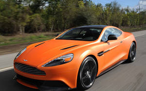 橙色Aston Martin在背景有樹木的道路上疾馳。 — 奧斯頓馬丁 壁紙