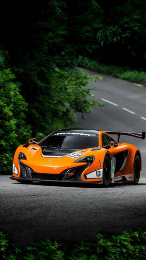 橙色McLaren賽車在森林道路上，周圍有綠樹。 — 汽車 iPhone 壁紙