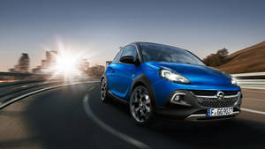 藍色Opel Adam Rocks在日落時分在蜿蜒的道路上行駛，背景是城市天際線。 — 歐寶 壁紙
