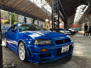 藍色日產Skyline R34停放在一個佈滿塗鴉的倉庫中，背景有人群。