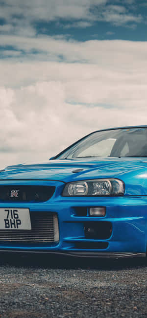 藍色日產Skyline R34跑車停放在礫石上，天空多雲。 — Pixel 11 Pro XL 壁紙
