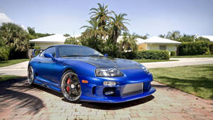 藍色跑車停在磚砌車道上，有棕櫚樹和房屋。