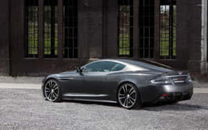啞光灰色 Aston Martin 跑車停泊在磚砌建築附近的碎石路上。 — Steam Deck OLED 壁紙