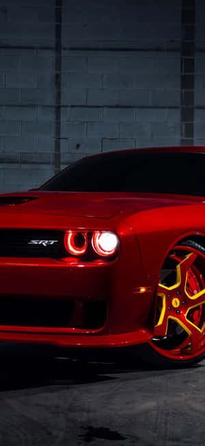 夜間停在磚牆前的紅色Dodge Challenger SRT，黃色輪圈。 — iPhone 16 Plus 壁紙