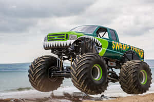 名為 Swamp Thing 的綠色怪獸卡車在海灘上靠近海洋跳躍。 — 卡車 壁紙