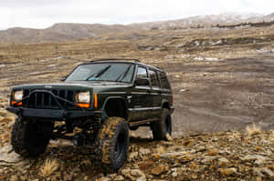 綠色Jeep Cherokee在岩石山地地形上行駛，背景是白雪覆蓋的山峰。 — MacBook Pro 14 壁紙