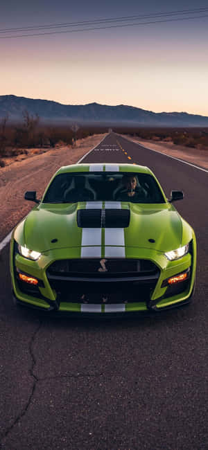 綠色Shelby Mustang在日落時分行駛在沙漠公路上。 — iPhone 14 Pro Max 壁紙