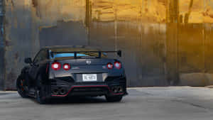 黑色日產GT-R R35跑車帶紅色飾邊，在金色光線下停靠在生鏽的工業牆邊。 — 日產GTR 壁紙