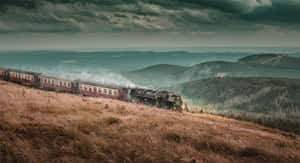 一輛蒸汽火車在多雲的天空下，沿著山坡穿過森林景觀行駛。 — 霍格華茲 GIF 壁紙