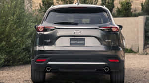 灰色Mazda CX-9 SUV後視圖，停在碎石上，靠近石牆和綠植。 — 馬自達 壁紙