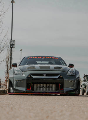 灰色日產GT-R賽車，帶有紅色飾邊，停泊在陰天下的瀝青路面上。 — 日產 壁紙