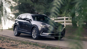 深灰色Mazda CX-9 SUV在鄉村道路上，周圍環繞著綠色樹葉和白色柵欄。 — 馬自達 壁紙