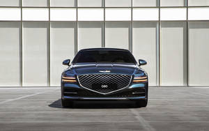 深藍色 Genesis G80 豪華轎車停放在現代建築物前的正面視圖。 — 創世紀 壁紙