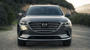灰色Mazda CX-9 SUV在日落時分靠近丘陵的碎石路上的正面視圖。 — 馬自達 壁紙