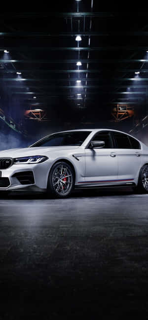 白色BMW M5跑車在光線昏暗的工業車庫中，配有聚光燈和煙霧。 — Galaxy A 壁紙