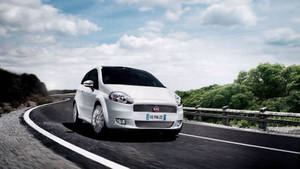白色Fiat Punto在晴朗的天空下在彎曲的道路上穿過綠蔭加速行駛。 — 飛雅特 壁紙