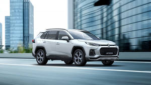 白色鈴木 Across SUV 快速穿過現代城市，玻璃建築在旁。 — 鈴木 壁紙