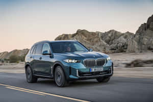藍綠色BMW X5在黃昏時分行駛在沙漠道路上，背景為岩石。 — MacBook Pro M4 壁紙